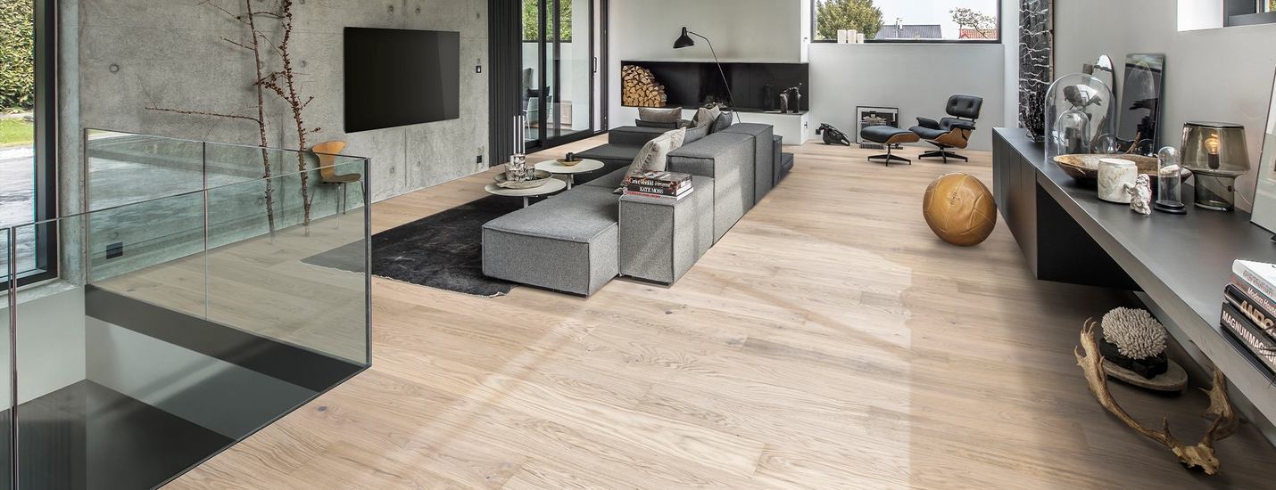 Modernes Wohnzimmer mit großen Fenstern, grauer Couchlandschaft und hellem Holzfußboden.