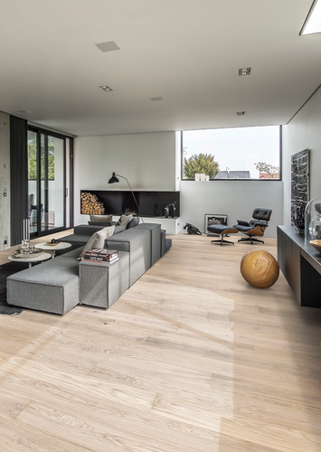 Modernes Wohnzimmer mit großen Fenstern, grauer Couchlandschaft und hellem Holzfußboden.