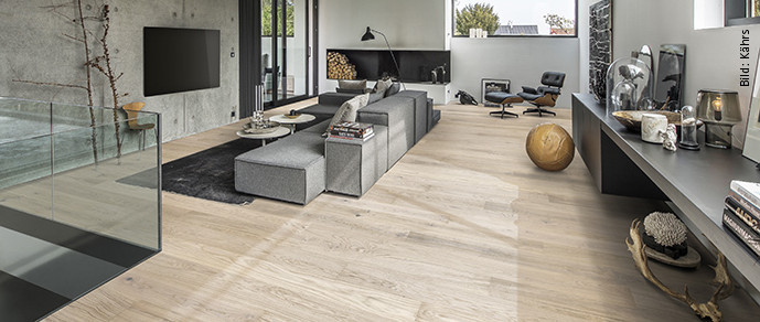 Modernes Wohnzimmer mit großen Fenstern, grauer Couchlandschaft und hellem Holzfußboden.