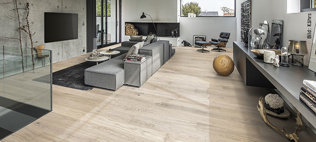 Modernes Wohnzimmer mit großen Fenstern, grauer Couchlandschaft und hellem Holzfußboden.