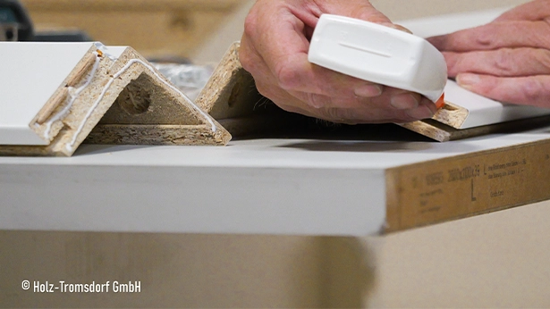Hand beim präzisen Auftragen von Klebstoff auf eine Holzleiste an der Werkbank, Detailaufnahme von Platten und Kanten. 