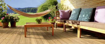 Gemütliche Holzterrasse mit Douglasien-Dielen, Lounge-Sofa mit Kissen, Holztisch, Hängematte und blühenden Pflanzen vor idyllischer Landschaft.