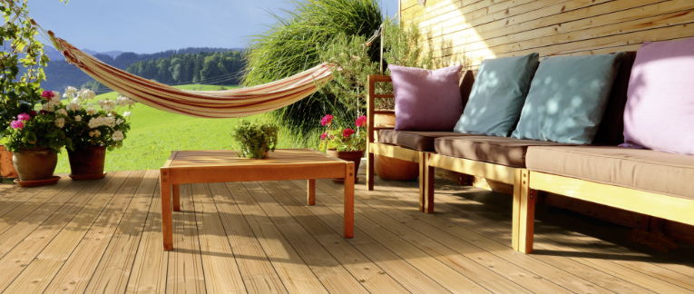 Gemütliche Holzterrasse mit Douglasien-Dielen, Lounge-Sofa mit Kissen, Holztisch, Hängematte und blühenden Pflanzen vor idyllischer Landschaft.