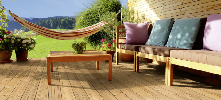 Gemütliche Holzterrasse mit Douglasien-Dielen, Lounge-Sofa mit Kissen, Holztisch, Hängematte und blühenden Pflanzen vor idyllischer Landschaft.